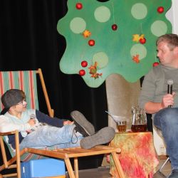 Ein Kind liegt auf einem Sommerstuhl mit einem Mikrofon in der Hand, ein Erwachsener sitzt auf einem Klapp Hocker mit Zeitung und Mikrofon in der Hand.