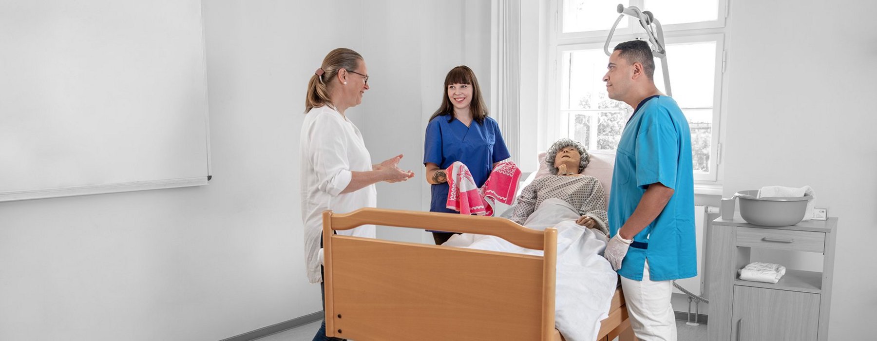Zwei Pflege auszubildende stehen vor einem Pflegedummy der im Bett liegt, während eine Lehrerin davor steht und etwas erklärt.