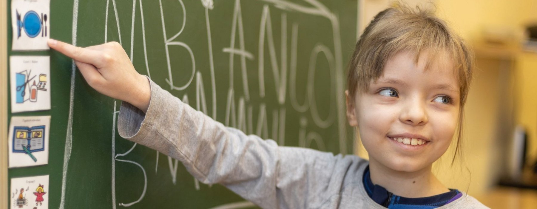Kleines Mädchen steht vor der Tafel und Zeigt auf ein Symbolbild mit einem Teller und Besteck