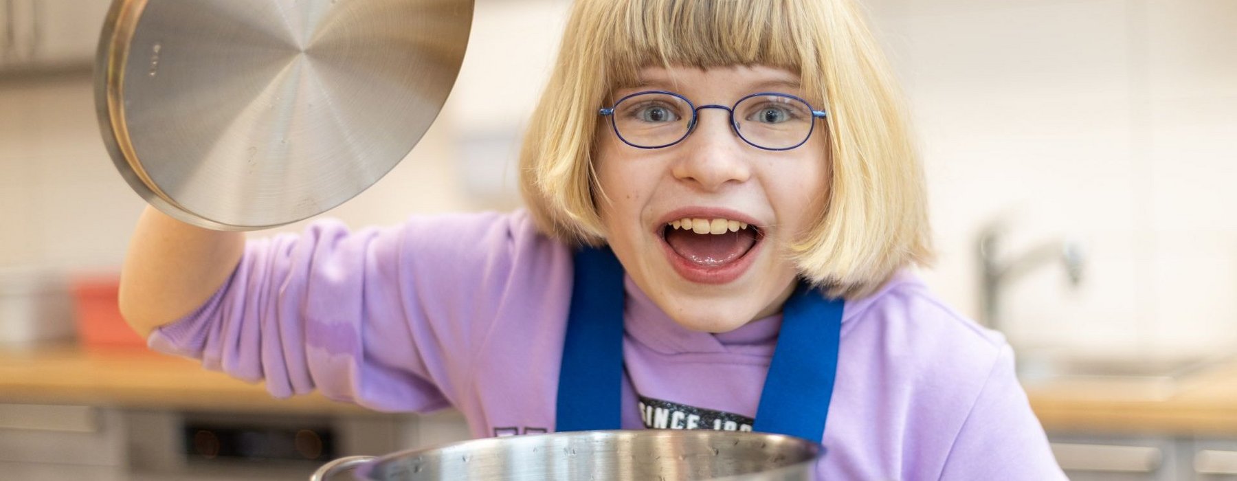 Mädchen mit Brille und blondem Bob schnitt öffnet voller Freude den Deckel eines Topfes