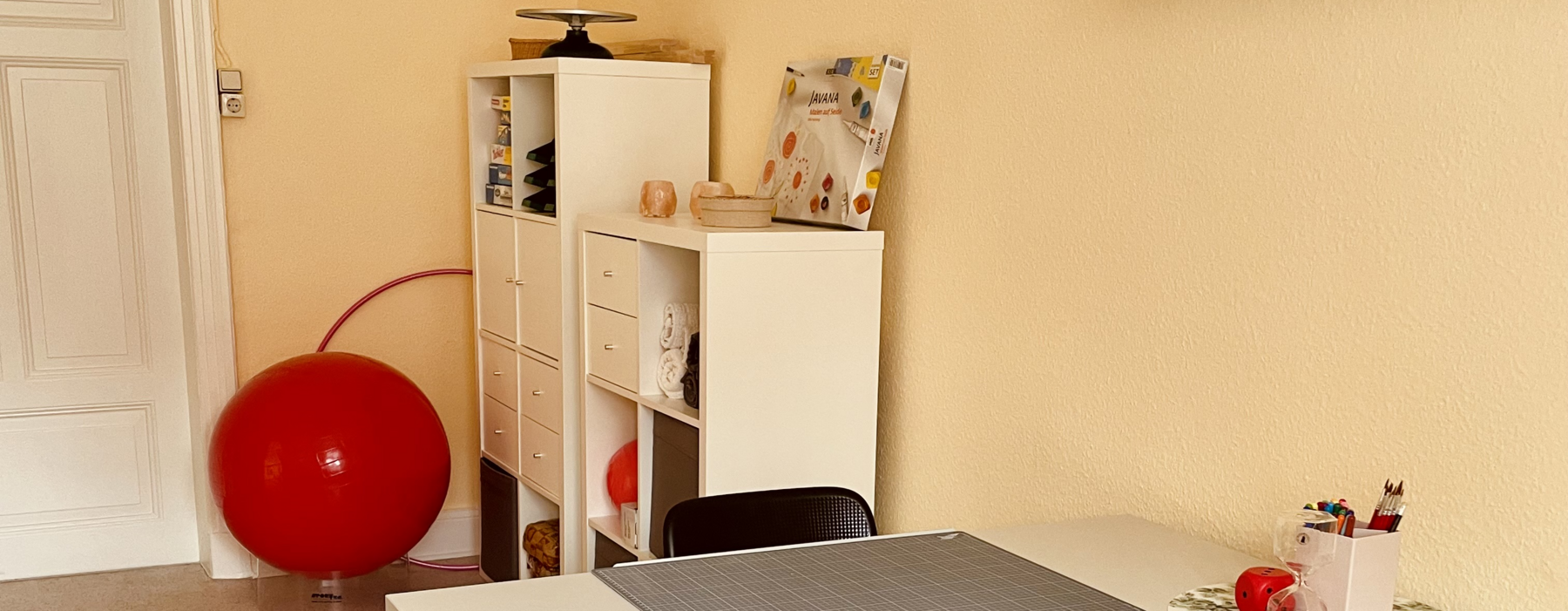 Hinter einem Tisch Stehen an der Wand ein Paar Regale und ein Gymnastik Ball.